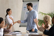 © fizkes - Smiling young businessman handshake excited female intern greeting with promotion, diverse colleagues applaud, happy male boss shake hand of employee congratulate with employment or work achievements