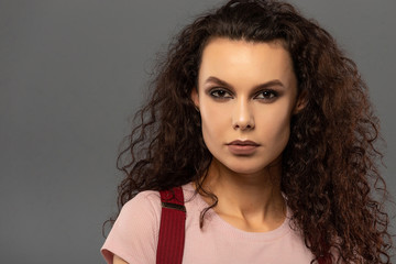 Young beautiful girl with curls on her head with a serious facial expression on a gray background. Portrait of a woman with a strong face with brown eyes in a pink T-shirt and red suspenders.