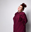 © FAB.1 - large red-haired model with a bun-shaped hairstyle, a dark pink tight-fitting striped dress and gold earrings. She is posing on a white background. The body is positive.
