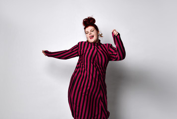 Wall Mural - Obese redhead lady in striped dress and earrings. She reaching her hand to you while posing sideways isolated on white background