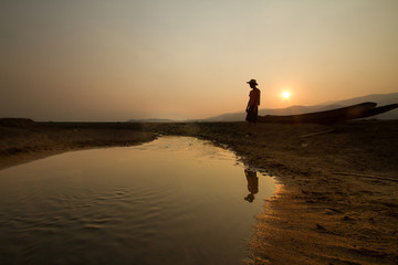 Sticker - Young man standing near a drying river with a wooden boat at the place that once had been a big lake before, climate change, water crisis concept.