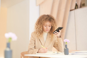 Wall Mural - Warm-toned portrait of curly-haired young woman using smartphone while working or studying sitting at table in cafe, copy space