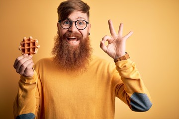 Redhead Irish man with beard eating sweet belgian waffle pastry over yellow background doing ok sign with fingers, excellent symbol