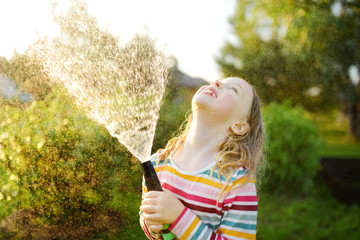 Wall Mural - Adorable girl playing with a garden hose on warm summer day. Child having fun with water on hot day. Outdoor summer activities for kids.