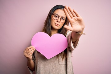 Wall Mural - Young beautiful brunette romantic woman holding big heart paper celebrating valentine day with open hand doing stop sign with serious and confident expression, defense gesture