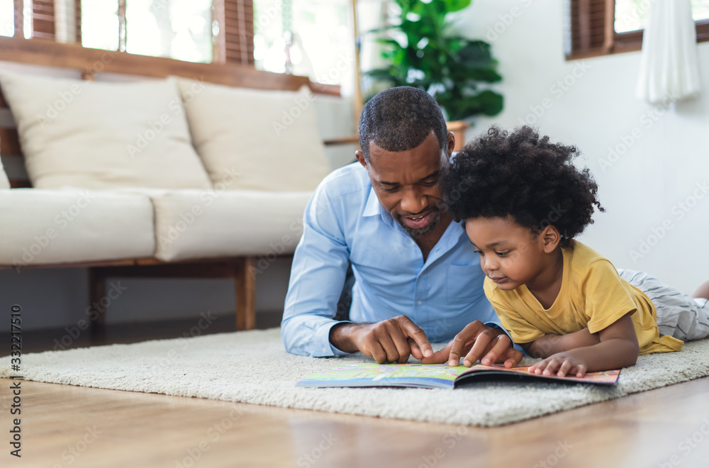 Happy African American father and son are reading a book and smiling while lying on floor spending time together at home. Children education and development concept. - obrazy, fototapety, plakaty 