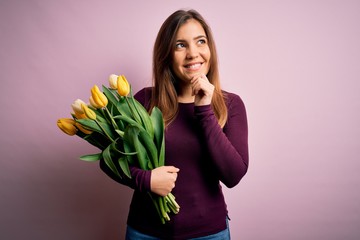 Sticker - Young blonde woman holding romantic bouquet of yellow tulips flowers over pink background with hand on chin thinking about question, pensive expression. Smiling and thoughtful face. Doubt concept.