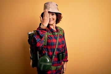 Poster - Middle age curly hair hiker woman hiking wearing backpack and water canteen using binoculars covering one eye with hand, confident smile on face and surprise emotion.