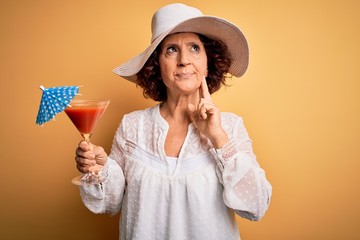 Poster - Middle age curly hair woman on holiday drinking cocktail over isolated yellow background serious face thinking about question, very confused idea