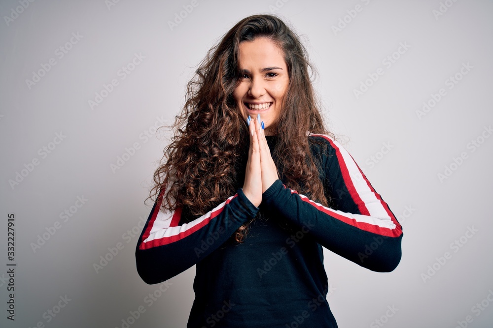 Young beautiful woman with curly hair wearing casual sweater over isolated white background praying with hands together asking for forgiveness smiling confident.