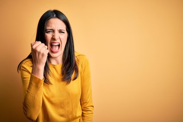 Wall Mural - Young brunette woman with blue eyes wearing casual sweater over yellow background angry and mad raising fist frustrated and furious while shouting with anger. Rage and aggressive concept.