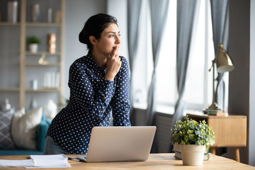 Wall Mural - Thoughtful Indian woman standing at desk, businesswoman freelancer pondering difficult task, looking out window, touching chin, pensive young female student working on research project
