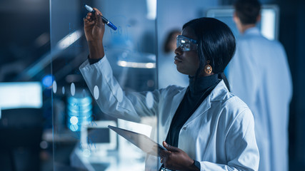 Wall Mural - In the Research Laboratory Smart and Beautiful African American Female Scientist Wearing White Coat and Protective Glasses Writes Formula on Glass Whiteboard, References Her Tablet Computer