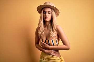 Poster - Young beautiful blonde woman on vacation wearing bikini and hat over yellow background with hand on stomach because indigestion, painful illness feeling unwell. Ache concept.