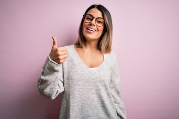 Young beautiful brunette woman wearing casual sweater and glasses over pink background doing happy thumbs up gesture with hand. Approving expression looking at the camera showing success.