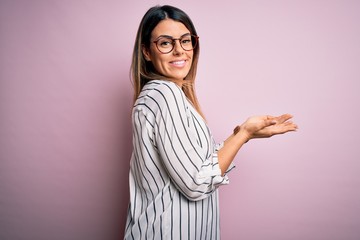 Sticker - Young beautiful woman wearing casual striped t-shirt and glasses over pink background pointing aside with hands open palms showing copy space, presenting advertisement smiling excited happy