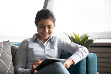 Smiling Indian woman using computer tablet, sitting on cozy couch in living room, happy girl looking at screen, watching funny video, chatting in social network or shopping online, playing game