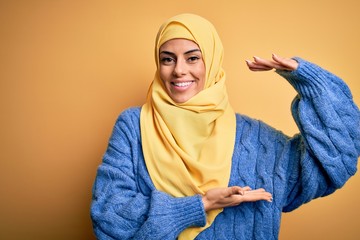 Poster - Young beautiful brunette muslim woman wearing arab hijab over isolated yellow background gesturing with hands showing big and large size sign, measure symbol. Smiling looking at the camera. Measuring