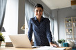 Portrait smiling Indian girl standing at desk with laptop, looking at camera, successful businesswoman freelancer posing for photo at workplace, satisfied excited female student in living room