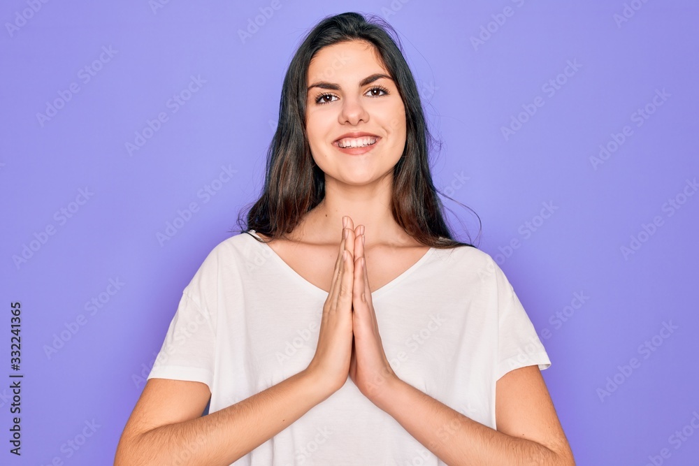 Young beautiful brunette woman wearing casual white t-shirt over purple background praying with hands together asking for forgiveness smiling confident.