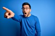 © Krakenimages.com - Young handsome man with beard wearing casual sweater and glasses over blue background Pointing with finger surprised ahead, open mouth amazed expression, something on the front