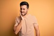 Young handsome man with beard wearing casual striped t-shirt over yellow background touching mouth with hand with painful expression because of toothache or dental illness on teeth. Dentist