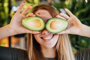 Happy young woman holding avocado halves under eyes