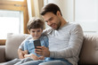 © fizkes - Happy young father sit relax on couch in living room with little son make selfie on smartphone, dad rest on sofa at home with small boy child watch video on cellphone enjoy leisure weekend together