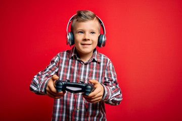 Adorable blond gamer toddler smiling happy and confident. Standing with smile on face playing video game using gamepad and headphones over isolated red background