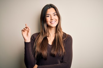 Young beautiful girl wearing casual sweater standing over isolated white background showing and pointing up with finger number one while smiling confident and happy.