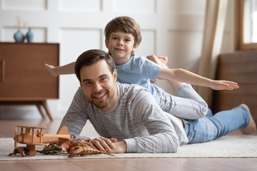 Wall Mural - Portrait of cute little boy lying with smiling young father on floor engaged in funny game together, happy small preschooler son playing with loving dad, enjoying family leisure weekend at home