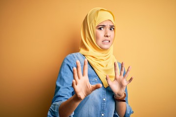 Poster - Young beautiful girl wearing muslim hijab standing over isolated yellow background afraid and terrified with fear expression stop gesture with hands, shouting in shock. Panic concept.