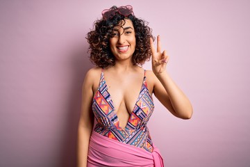 Sticker - Young beautiful arab woman on vacation wearing swimsuit and sunglasses over pink background smiling with happy face winking at the camera doing victory sign. Number two.