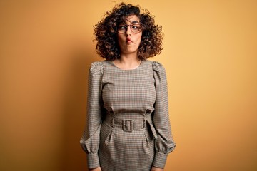 Beautiful arab business woman wearing dress and glasses standing over yellow background making fish face with lips, crazy and comical gesture. Funny expression.