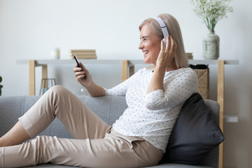 Side view happy middle aged woman enjoying favorite music in wireless headphones, relaxing on comfortable couch at living room. Smiling satisfied female retiree choosing songs of youth in mobile app.