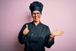 Middle age brunette chef woman wearing cooker uniform and hat over isolated pink background Showing palm hand and doing ok gesture with thumbs up, smiling happy and cheerful