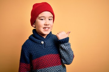 Sticker - Young little boy kid wearing wool cap and winter sweater over yellow isolated background smiling with happy face looking and pointing to the side with thumb up.