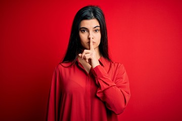 Wall Mural - Young beautiful brunette woman wearing casual shirt standing over red background asking to be quiet with finger on lips. Silence and secret concept.