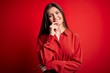 © Krakenimages.com - Young beautiful woman with blue eyes wearing casual shirt standing over red background looking confident at the camera with smile with crossed arms and hand raised on chin. Thinking positive.
