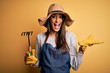 © Krakenimages.com - Young beautiful farmer woman wearing apron and hat using rake over yellow background very happy and excited, winner expression celebrating victory screaming with big smile and raised hands