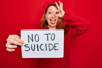 Canvas Print - Beautiful redhead woman holding banner showing no to suicide message over red background with happy face smiling doing ok sign with hand on eye looking through fingers