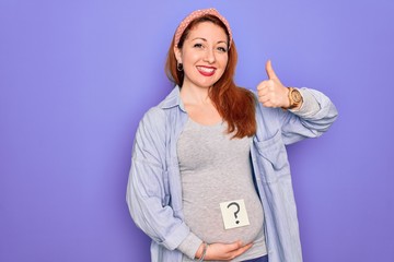 Wall Mural - Young beautiful redhead pregnant woman expecting baby with question mark on stomach happy with big smile doing ok sign, thumb up with fingers, excellent sign