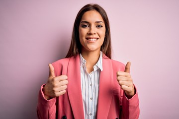 Young beautiful brunette woman wearing jacket standing over isolated pink background success sign doing positive gesture with hand, thumbs up smiling and happy. Cheerful expression and winner gesture.