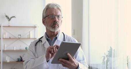 Wall Mural - Serious senior adult male doctor using digital tablet at work. Elderly old professional physician reading online news, checking medical healthcare technology data on pad computer in hospital.