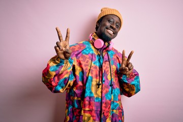 Poster - young handsome african american man wearing colorful coat and cap over pink background smiling looki