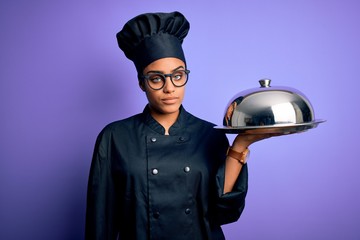 Canvas Print - Young african american cooker girl wearing uniform and hat holding tray with dome with a confident expression on smart face thinking serious