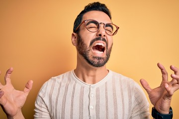 Sticker - Young handsome man with beard wearing casual t-shirt and glasses over yellow background crazy and mad shouting and yelling with aggressive expression and arms raised. Frustration concept.