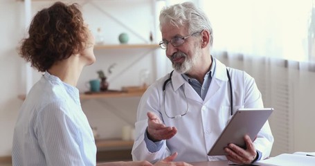 Wall Mural - Smiling senior doctor showing good medical test results on tablet handshaking female patient at hospital checkup. Happy old elder doc and healthy recovered woman client shake hands. Gratitude concept.