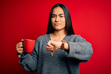 Canvas Print - Young beautiful asian woman drinking mug of coffee standing over isolated red background with angry face, negative sign showing dislike with thumbs down, rejection concept