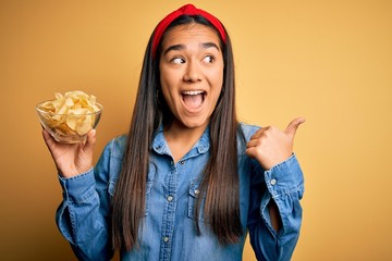 Canvas Print - Young beautiful asian woman holding bowl with chips potatoes over isolated yellow background pointing and showing with thumb up to the side with happy face smiling